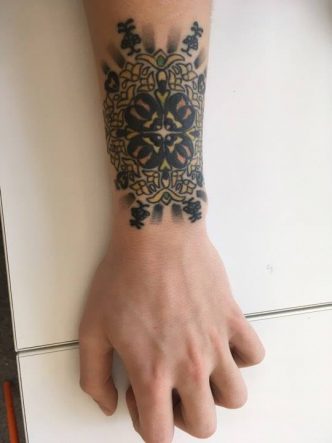 Image of gold and yellow coloured mandala tattoo with black shading on caucasian man's left outer wrist.
